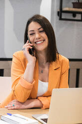 Smiling female freelancer sitting at table with laptop and using a phone - ADSF28992