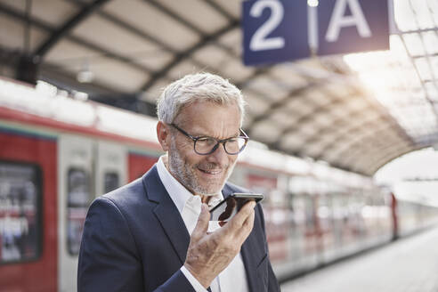 Lächelnder Geschäftsmann, der eine Sprachnachricht über ein Mobiltelefon am Bahnsteig sendet - RORF02833