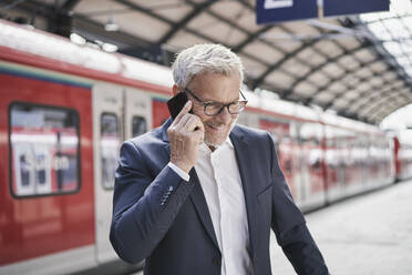 Lächelnder männlicher Berufstätiger, der am Bahnhof mit einem Mobiltelefon spricht - RORF02835