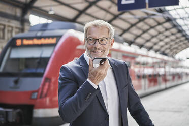 Lächelnder männlicher Berufstätiger, der wegschaut, während er eine Voicemail über ein Smartphone auf einem Bahnhof sendet - RORF02836