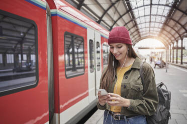 Junger weiblicher Fahrgast, der im Zug sein Smartphone benutzt und über Kopfhörer Musik hört - RORF02843