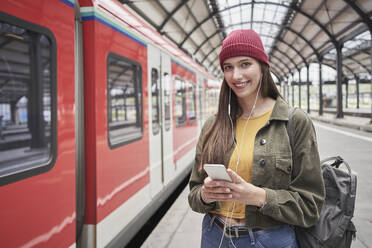 Beautiful female passenger with mobile phone smiling while standing by train at railroad station - RORF02845