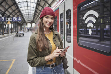 Lächelnder junger weiblicher Fahrgast mit Smartphone, der auf dem Bahnhof Musik über sein Smartphone hört - RORF02847