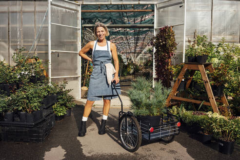 Mature female farmer with cart standing at greenhouse doorway - VPIF04648