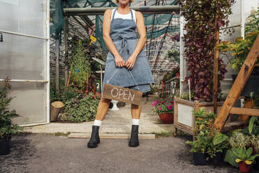 Female greenhouse worker holding wooden open sign at doorway of greenhouse - VPIF04675