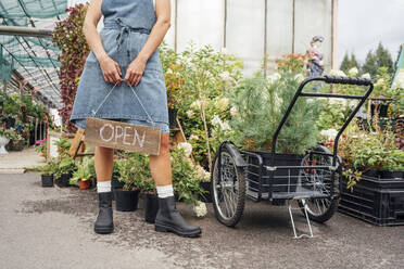 Female greenhouse worker holding open sign while standing by plants in greenhouse - VPIF04676