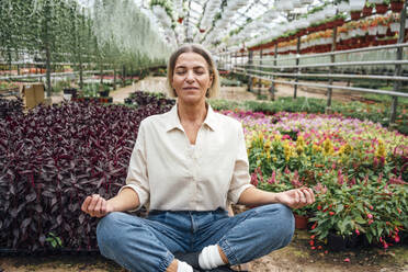 Female farmer practicing yoga while sitting cross-legged in greenhouse - VPIF04705