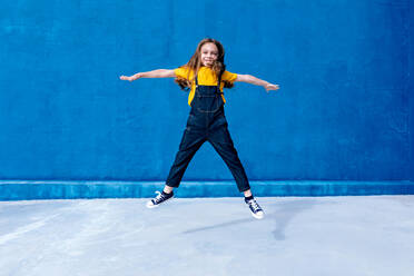 Carefree smiling teenager in moment of jumping above ground with outstretched arms on blue background - ADSF29042