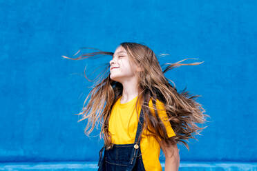 Entzückter verträumter Teenager mit langen fliegenden Haaren stehend mit geschlossenen Augen auf blauem Hintergrund - ADSF29046