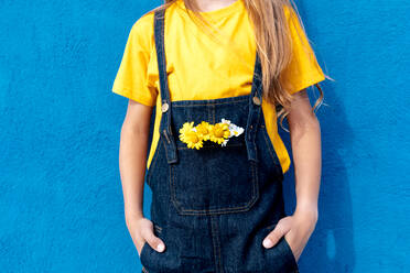 Unkenntlich gemachter Teenager-Hipster mit einem gelben Blumenstrauß in der Tasche seines Jeans-Overalls, der an einer blauen Wand lehnt - ADSF29049