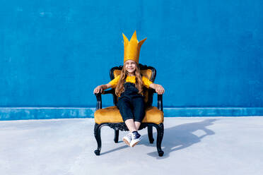 Zufriedener Teenager mit goldener Papierkrone, der wie ein König auf einem Thron sitzt, auf blauem Hintergrund und in die Kamera schaut - ADSF29052