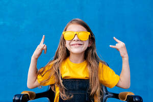 Ausdrucksstarker Teenager mit langen Haaren und trendiger gelber Sonnenbrille, der ein Rockschild zeigt und auf blauem Hintergrund schreit - ADSF29055