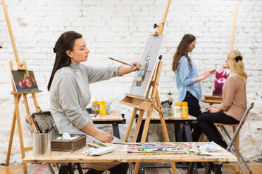 Seitenansicht einer fokussierten Künstlerin, die auf einer Staffelei in einem Kunstatelier auf Leinwand malt, vor einem Hintergrund von unscharfen Frauen - ADSF29061