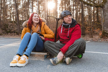 Vergnügtes Paar auf Skateboard und Roller sitzend, während es auf einem Parkplatz im Herbst Spaß hat - ADSF29062