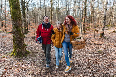 Lächelndes Paar und Tochter im Teenageralter, die mit Korb und Plaid im Wald spazieren gehen, um ein Picknick im Herbst zu machen - ADSF29066