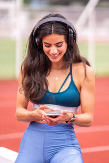 Cheerful young athletic Hispanic female in sportswear and headphones listening to music and browsing mobile phone while having break after training on stadium track - ADSF29072