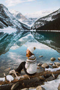 Rückenansicht einer nachdenklichen Touristin in Pullover und Hut, die mit geschlossenen Augen am Ufer des Lake Louise vor einem verschneiten Bergkamm an einem Wintertag in Alberta, Kanada, sitzt - ADSF29113
