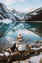 Rückenansicht einer nachdenklichen anonymen Touristin in Pullover und Hut, die am Ufer des Lake Louise vor einem verschneiten Bergkamm an einem Wintertag in Alberta, Kanada, sitzt - ADSF29114