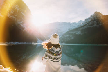 Rückenansicht einer anonymen Wanderin in warmer Kleidung, die an einem sonnigen Wintermorgen im Banff National Park gegen den ruhigen Lake Louise und die stehenden Berge läuft - ADSF29116