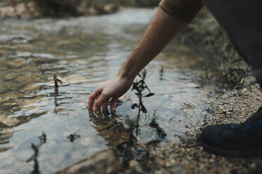 Seitenansicht eines anonymen männlichen Reisenden, der seine Hand in das saubere Wasser eines seichten Baches hält, während er sich nach einer Wanderung in der Natur ausruht - ADSF29120