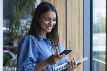 Smiling female freelancer using mobile phone while standing in coffee shop - PNAF02127