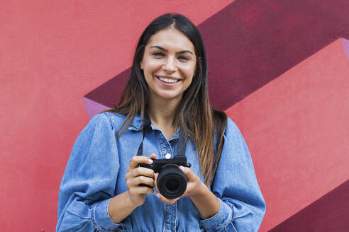 Lächelnde freiberufliche Mitarbeiterin mit Kamera vor einer Wand stehend - PNAF02132