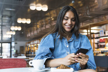 Lächelnde Geschäftsfrau, die ein Mobiltelefon benutzt, während sie in einem Café sitzt - PNAF02133