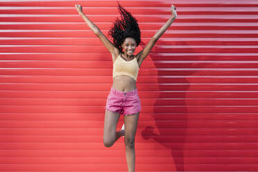 Happy young woman with arms raised jumping in front of red shutter - JRVF01634