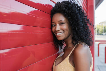 Lächelnde junge Frau mit lockigem Haar lehnt sich an einen roten Fensterladen - JRVF01637