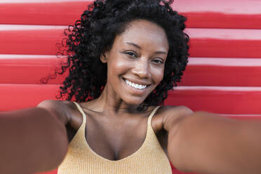 Lächelnde junge Afro-Frau mit lockigem Haar vor einem roten Fensterladen stehend - JRVF01642