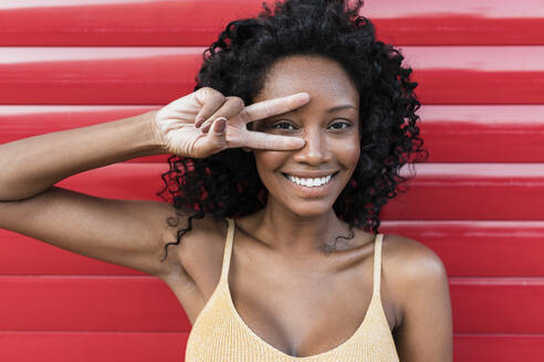 Lächelnde Frau mit lockigem Haar, die vor einem roten Fensterladen ein Friedenszeichen macht - JRVF01653