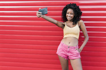 Cheerful woman with hands in pockets taking selfie through smart phone in front of red shutter - JRVF01661