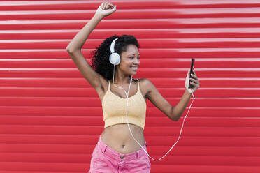 Happy woman with hand raised dancing while listening music through headphones in front of red shutter - JRVF01663