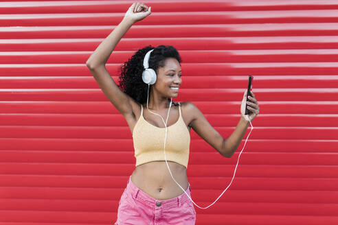 Glückliche Frau mit erhobener Hand, die tanzt, während sie Musik über Kopfhörer vor einem roten Fensterladen hört - JRVF01663
