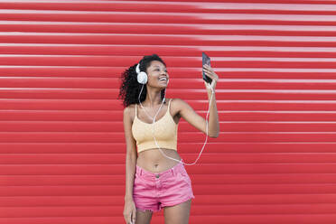 Fröhliche junge Frau, die ihr Smartphone in der Hand hält und über Kopfhörer Musik hört, vor einem roten Fensterladen - JRVF01664