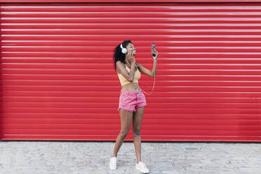 Lächelnde junge Frau, die ein Selfie mit ihrem Smartphone macht, während sie vor einem roten Fensterladen Musik hört - JRVF01667