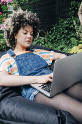 Woman using laptop while relaxing on bean bag at backyard - ASGF01230