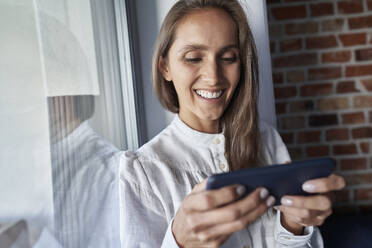 Lächelnde Frau, die ihr Smartphone bei einem Glas zu Hause benutzt - ABIF01469