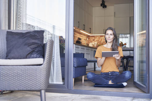 Nachdenkliche Frau mit digitalem Tablet, die in der Nähe einer Tür sitzt - ABIF01474