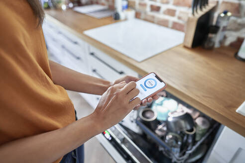 Woman operating dishwasher through mobile application at modern home - ABIF01496