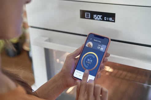 Woman adjusting oven temperature on smart phone at modern home - ABIF01498