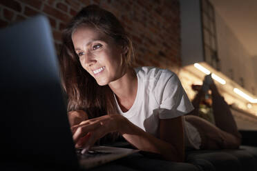 Junge Frau, die einen Laptop benutzt, während sie nachts auf dem Sofa zu Hause liegt - ABIF01513