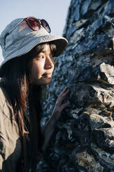 Young woman looking away while standing behind rock on sunny day - ASGF01246