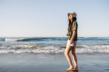 Junge Frau mit Hut steht am Strand an einem sonnigen Tag - ASGF01250