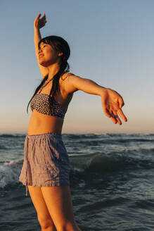 Unbekümmerte junge Frau mit ausgestreckten Armen am Strand stehend - ASGF01260