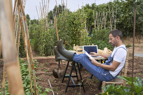 Male farmer reading clipboard while sitting with feet up at table in agricultural field - VEGF04917