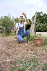 Smiling farm worker holding garden hoe and potato while working in agricultural field - VEGF04925