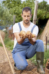 Male farmer holding potatoes while crouching in agricultural field - VEGF04926