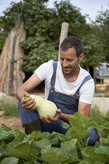 Lächelnder männlicher Landwirt hält Kürbis, während er auf einem landwirtschaftlichen Feld hockt - VEGF04933
