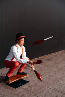 Ganzer Körper eines agilen jungen männlichen Zirkuskünstlers, der mit Keulen jongliert und auf einem Brett balanciert - ADSF29138